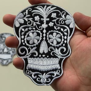 May include: Embroidered black skull patch with intricate white floral and scrollwork details. The patch features a detailed sugar skull design, perfect for adding to clothing or accessories. The patch is approximately 4 inches wide.