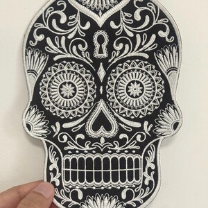 May include: Black and white embroidered skull patch with intricate floral and keyhole designs. The patch features detailed stitching, resembling a sugar skull, and is likely for decorative purposes.