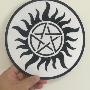 Op de afbeelding: Een ronde, witte patch met een zwarte rand. Het ontwerp toont een zwart pentagram in een cirkel, omringd door gestileerde zwarte vlammen. De patch wordt tegen een effen witte achtergrond gehouden.