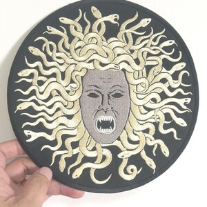 Puede incluir: Un parche redondo y negro con una imagen bordada de la cara de Medusa. La cara es gris con la boca abierta y dientes afilados. Su cabello es una masa de serpientes beige. El parche está sostenido por una mano.