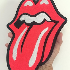 May include: A large, embroidered patch featuring the iconic Rolling Stones tongue and lips logo. The design has a bright red tongue with white teeth and a black outline. The patch is held up against a white background.