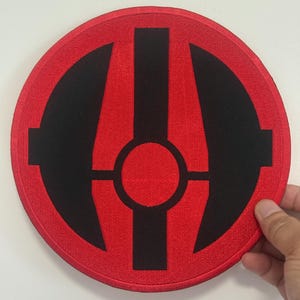 Może przedstawiać: Okrągła, czerwona naszywka z czarnym geometrycznym wzorem. Wzór przedstawia centralne koło z pionową linią wychodzącą z niego, przeciętą przez linie poziome. Naszywka jest trzymana przez dłoń.