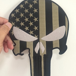 May include: Embroidered patch in the shape of a skull, featuring the American flag design. The top left section has stars, while the right side has vertical stripes. The skull's eyes and teeth are in a contrasting color.