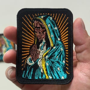 May include: Embroidered patch depicting a skeletal figure in a blue and yellow hooded cloak, with hands clasped in prayer. The design includes gold rays emanating from behind the figure, set against a black background.