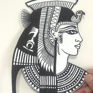 Könnte beinhalten: Schwarz-weißer Aufnäher mit dem Profil einer ägyptischen Königin. Das Design umfasst einen detaillierten Kopfschmuck, einen aufwendigen Kragen und Hieroglyphen. Der Aufnäher wird von einer Hand gehalten.