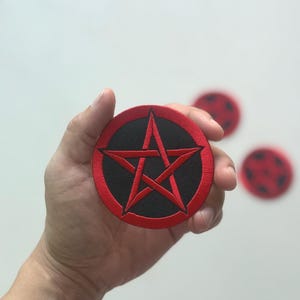 May include: Embroidered patch featuring a red pentagram within a black circle, surrounded by a red border. The patch is round and has a textured appearance. Additional patches are visible in the background.