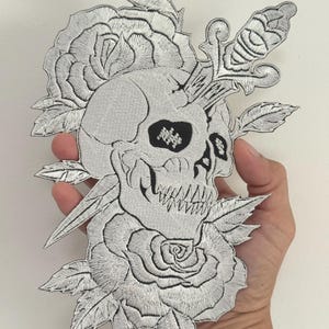 May include: Embroidered patch featuring a skull pierced by a dagger, surrounded by roses and leaves. The design is in shades of gray and white, with detailed stitching. A gothic-themed patch.