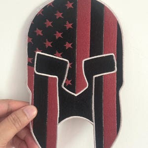 Peut inclure: Un écusson de casque spartiate noir et rouge avec un motif rappelant le drapeau américain. Le coin supérieur gauche présente des étoiles rouges sur fond noir, tandis que le reste présente des rayures verticales rouges et noires. L'écusson est tenu par une main.