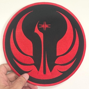 May include: A round, black patch with a red emblem. The emblem features a stylized, symmetrical design with a central vertical element and wing-like shapes extending outwards. The patch is held by a hand.