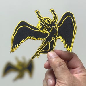 Könnte beinhalten: Schwarz-goldener, gestickter Aufnäher mit einer geflügelten Figur mit erhobenen Armen. Der Aufnäher hat ein detailliertes Design mit schwarzem Hintergrund und goldener Umrandung. Der Aufnäher wird von einer Hand gehalten.