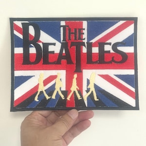 Könnte beinhalten: Rechteckiger Aufnäher mit dem Design der britischen Flagge und dem Schriftzug "THE BEATLES" in schwarzen Blockbuchstaben. Darunter vier gelbe Silhouetten von Personen, die gehen. Der Aufnäher hat einen schwarzen Rand.