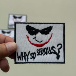 May include: Square white patch with a black border featuring a stylized image of a smiling face with black eye mask and red lips. The phrase "WHY SO SERIOUS?" is embroidered in black.