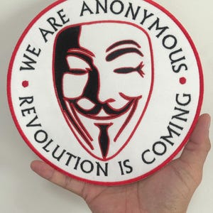 May include: Round white patch with a red border featuring the Anonymous mask in black and red. The text "WE ARE ANONYMOUS" and "REVOLUTION IS COMING" are in black, encircling the mask.