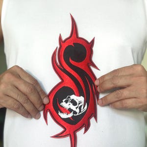Può includere: Toppa ricamata con un design rosso e nero e un motivo a teschio. La toppa ha una forma distintiva con bordi affilati e appuntiti. Il teschio è bianco e dettagliato, su sfondo nero.