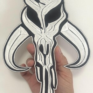 May include: Embroidered patch with a black and white skull design and curved horns. The patch has a detailed, textured appearance with a white outline and black fill. The patch is held in a hand.