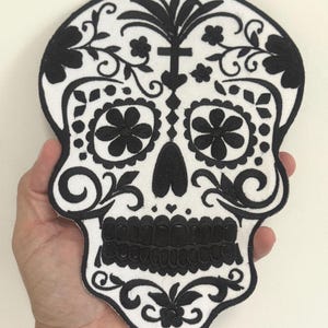 May include: A black and white embroidered skull patch with floral and ornamental details. The patch features a skull shape with intricate designs, including flowers, swirls, and a cross-like element. The design is set against a white background.