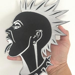 May include: A black and silver embroidered patch featuring a punk rock profile with a mohawk, tongue sticking out, and a studded collar. The patch is held up against a white background.