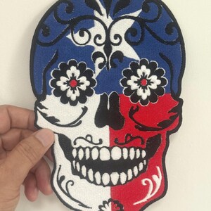 May include: Embroidered skull patch with a blue, white, and red design. The top half is blue with white star and floral accents. The bottom half is split between white and red, with black floral accents and a detailed toothy grin.