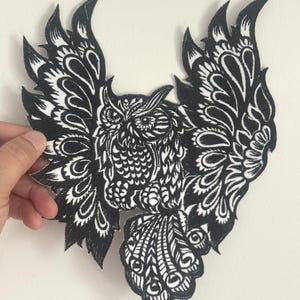 以下が含まれることがあります： 黒と白の刺繍ワッペン、翼を広げたフクロウ。 羽や顔の特徴など、複雑なディテールが特徴です。 フクロウは、ジャケット、バッグ、その他の布地アイテムに適した様式化されたデザインです。