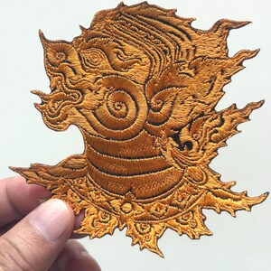 May include: A detailed, gold-colored embroidered patch featuring a stylized mythical creature's head. The patch has intricate patterns and textures, with a spiky, ornate design. The patch is held by a hand, showcasing the detailed craftsmanship.