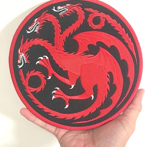 May include: A round, embroidered patch featuring a red three-headed dragon emblem on a black background. The dragon has detailed wings and sharp claws. The patch is held in a hand against a white background.