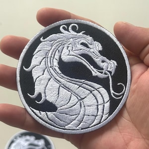 Peut inclure: Écusson brodé circulaire avec un motif de dragon blanc sur fond noir. Le dragon est très détaillé. L'écusson est tenu dans une main, avec un autre écusson visible en arrière-plan.