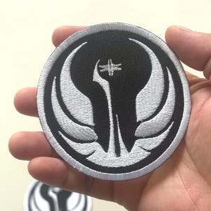 Op de afbeelding: Een ronde, zwart-witte geborduurde patch met een gestileerd embleem. Het ontwerp bevat een centraal verticaal element met een kruisvorm aan de bovenkant, geflankeerd door gebogen, vleugelachtige vormen. De patch wordt in een hand gehouden.