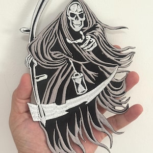 May include: Embroidered patch featuring the Grim Reaper in black and gray. The Reaper is depicted with a skull face, hooded cloak, and scythe. A small hourglass is visible. The patch is held in a hand.