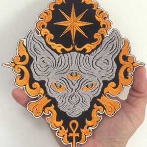 May include: Embroidered patch featuring a stylized cat face with three eyes, set against a black background with a gold star and ankh symbol. The cat is gray with orange and blue accents, framed by an ornate gold border.