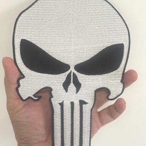 May include: A white and black skull patch with a detailed embroidered design. The skull features black eye sockets and a nose, with vertical lines forming the teeth. The patch is held in a hand.