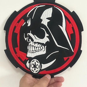 May include: Embroidered patch featuring a stylized skull design within a black and red circular frame. The design includes a skull-like helmet and a symbol. The patch is likely for clothing or accessories.