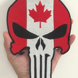 May include: Embroidered patch in the shape of a skull, featuring the Canadian flag design. The patch has a red, white, and black color scheme, with a maple leaf in the center. The skull's eyes are black.
