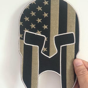 May include: Embroidered patch in the shape of a Spartan helmet, featuring a design that combines the American flag with the helmet's silhouette. The patch has black and tan stripes, with gold stars on a black background.