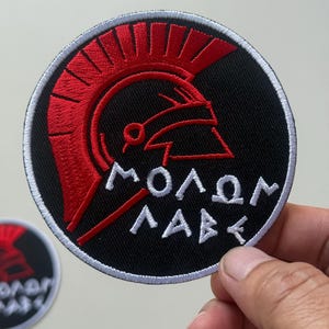 Puede incluir: Parche bordado circular negro con un casco espartano rojo. El parche incluye la frase griega "ΜΟΛΩΝ ΛΑΒΕ" en letras blancas, con un borde blanco.