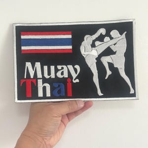 Peut inclure: Écusson rectangulaire noir avec les mots "Muay Thai" en blanc, rouge et bleu. L'écusson présente un drapeau thaïlandais et un graphique de deux figures engagées dans un combat d'arts martiaux. L'écusson a une bordure blanche.