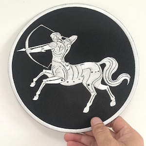 May include: A black round patch featuring a white embroidered Sagittarius design. The design depicts a centaur archer with a bow and arrow. The patch has a white border and is held by a hand.