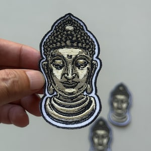 May include: Embroidered patch featuring a detailed Buddha head with a serene expression. The patch has a black border with white stitching, and the Buddha's face is in shades of gold and brown. Two smaller patches are also visible.