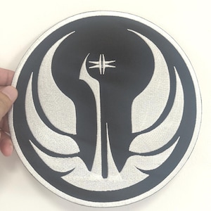 May include: A round, black patch with a white embroidered emblem. The emblem features a stylized design with wing-like shapes and a central vertical element. The patch is likely for clothing or accessories.