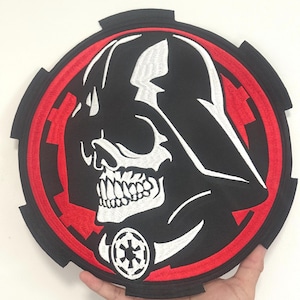 May include: A large, round black and red patch featuring a skull-like Darth Vader design. The patch has a white skull and the Imperial symbol. The patch is held by a hand.