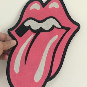Puede incluir: Un parche bordado rosa y negro con el icónico logotipo de la lengua y los labios de los Rolling Stones. La lengua es rosa claro con detalles blancos, y los labios están delineados en negro. El parche está sostenido por una mano sobre un fondo blanco.