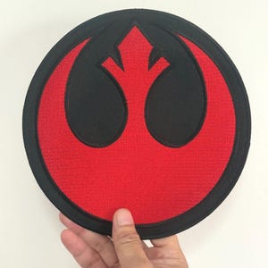 Puede incluir: Un parche redondo bordado con el símbolo de la Alianza Rebelde de Star Wars. El diseño presenta un emblema negro sobre un fondo rojo, con un borde negro. El parche se muestra sobre un fondo blanco.