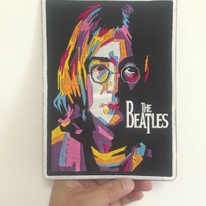 Könnte beinhalten: Gestickter Aufnäher mit einem farbenfrohen, geometrischen Porträt von John Lennon mit Brille. Der Aufnäher hat einen schwarzen Hintergrund und den Text "THE BEATLES" in Weiß. Der Aufnäher ist rechteckig mit silberner Umrandung.