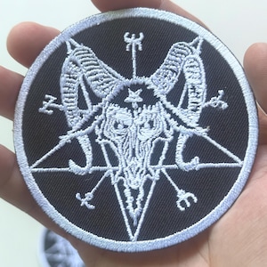 Pode incluir: Um patch bordado circular preto com um símbolo de Baphomet branco. O design inclui um pentagrama, uma cabeça de cabra e vários símbolos esotéricos. O patch é segurado em uma mão.