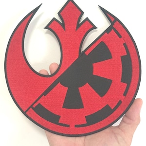 Op de afbeelding: Een rood en zwart geborduurd embleem met een gesplitst ontwerp. De ene helft toont een gestileerd embleem met een gebogen vorm, de andere een cirkelvormig, tandwielachtig ontwerp. Het embleem wordt in een hand gehouden.