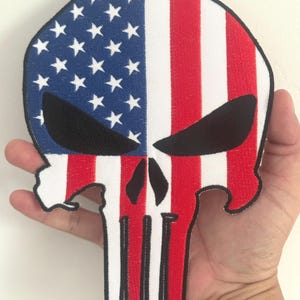 May include: Embroidered skull-shaped patch with an American flag design. The top section is blue with white stars, and the lower half has red and white stripes. The skull's eye sockets are black.