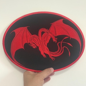 Puede incluir: Parche bordado con un dragón rojo con alas extendidas sobre un fondo ovalado negro. El dragón está intrincadamente detallado, con un cuerpo serpentino y grandes alas texturizadas. El parche tiene un borde rojo.