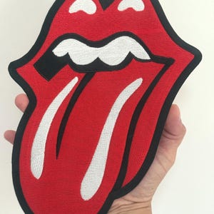 Könnte beinhalten: Gestickter Aufnäher mit dem berühmten roten Lippen- und Zungen-Logo mit schwarzem Rand. Die Zunge hat weiße Akzente. Der Aufnäher wird in einer Hand vor weißem Hintergrund gehalten. Ein Musik-Accessoire.