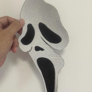 May include: A silver and black patch in the shape of the iconic Scream mask. The mask has a textured silver fabric with large black eye and mouth openings. The patch is held against a white background.