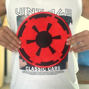 Può includere: Una canotta bianca con la scritta "VINTAGE" stampata sopra. Una grande toppa circolare ricamata rossa e nera con un design geometrico è centrata sulla maglietta. La maglietta ha anche le parole "CLASSIC CARS" stampate sopra.