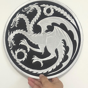 May include: A circular, black and silver embroidered patch with a three-headed dragon motif. The dragons are silver with detailed wings and scales, set against a black backdrop. The patch is round.